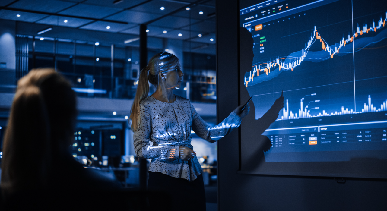A woman in an office setting gestures toward a large screen displaying dynamic financial charts